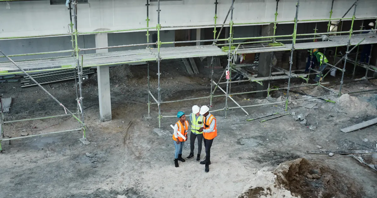Tres trabajadores con chalecos reflectantes y cascos revisan planos en una obra con andamios